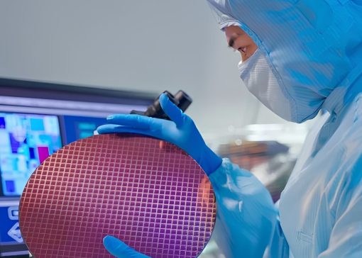 Scientist in full cleanroom gear examines a round semiconductor wafer near a computer monitor with test data in the background.