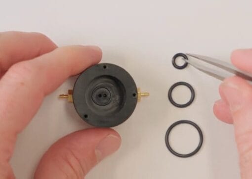 Close-up of hands disassembling a small circular device with O-rings, using tweezers to place a tiny seal.