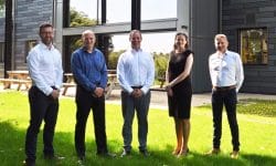 Five professionals (four men and one woman) stand in a row on a sunny grassy area outside a modern office building, posing for the photo.