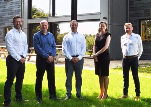 Five professionals (four men and one woman) stand in a row on a sunny grassy area outside a modern office building, posing for the photo.