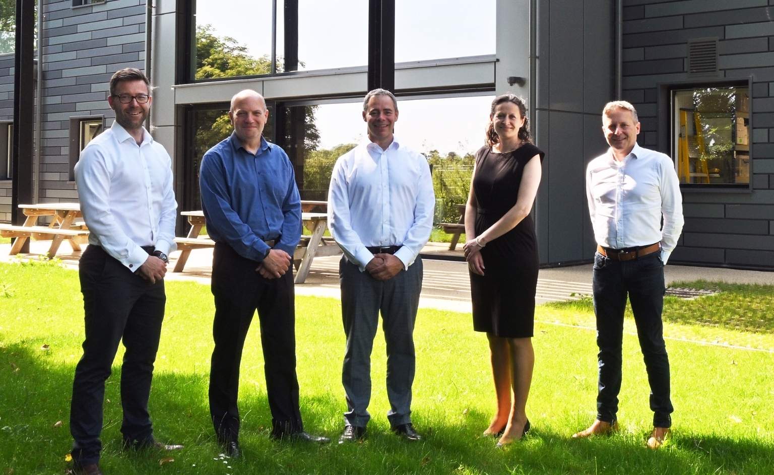 Board of Directors - web | ION SENSE Five professionals (four men and one woman) stand in a row on a sunny grassy area outside a modern office building, posing for the photo.