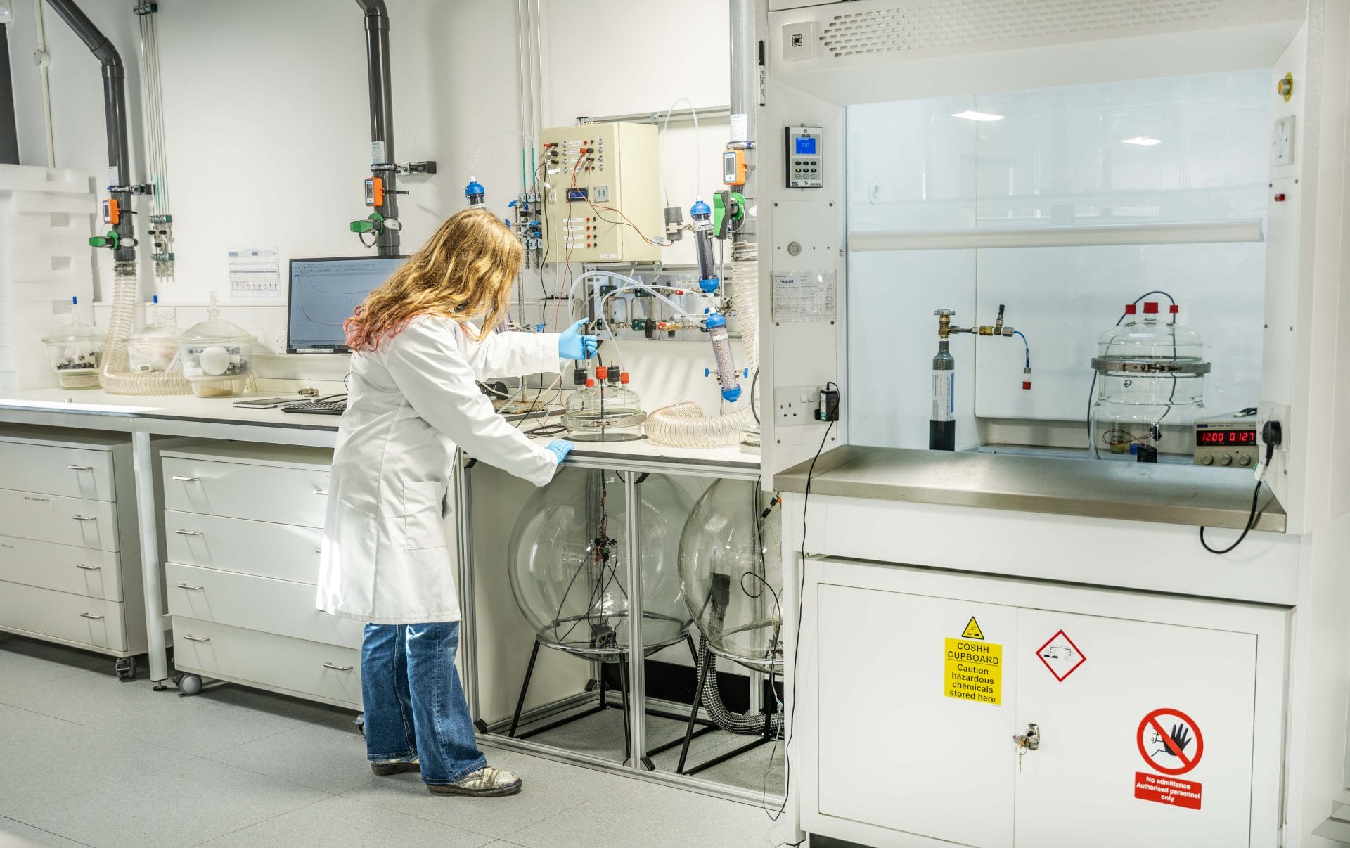 Laboratory technician in a white coat and blue gloves handling glassware and tubing at a workbench in a chemistry lab.