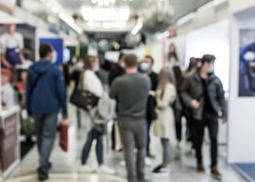 Crowded indoor exhibition hall with people walking and talking; several attendees wear face masks.
