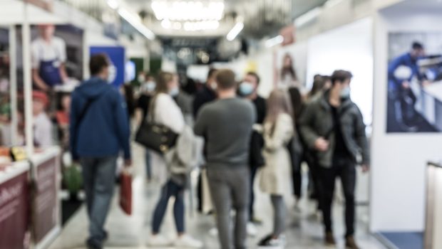 Crowded indoor exhibition hall with people walking and talking; several attendees wear face masks.