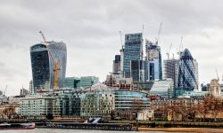 London skyline along the Thames with the Walkie Talkie building on the left, The Gherkin on the right, and construction cranes overhead under a cloudy sky.