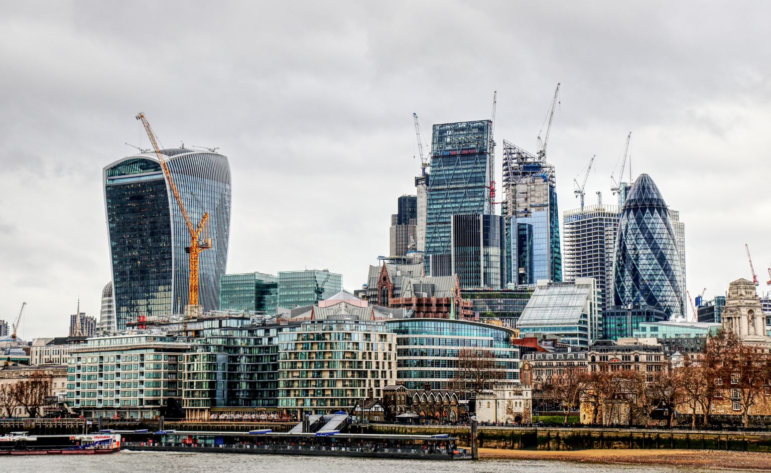 London skyline along the Thames with the Walkie Talkie building on the left, The Gherkin on the right, and construction cranes overhead under a cloudy sky.