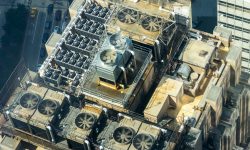 Aerial view of a building roof with numerous large ventilation fans and machinery arranged in rows under metal walkways.",