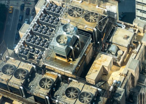 Aerial view of a building roof with numerous large ventilation fans and machinery arranged in rows under metal walkways.",