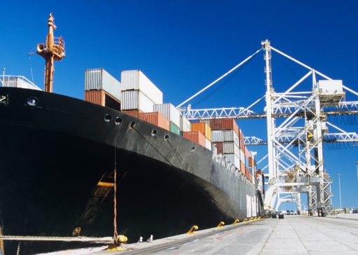 Large cargo ship docked at a port with stacked shipping containers and towering white gantry cranes against a clear blue sky.