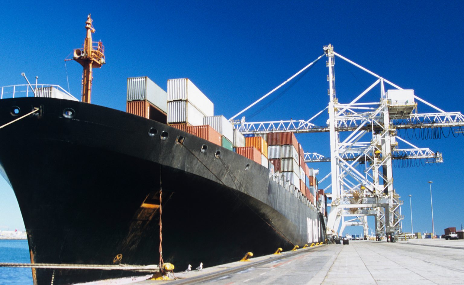 Real-Time VOC Detection at Harbours and Marine Terminals | ION SENSE Large cargo ship docked at a port with stacked shipping containers and towering white gantry cranes against a clear blue sky.